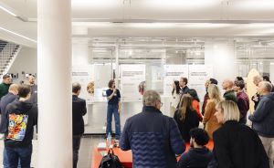 Eine Frau präsentiert vor einer Gruppe von Menschen in einem modernen, hellen Raum mit Ausstellungstafeln an der Wand.