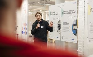 Mann mit Mikrofon spricht vor Ausstellungstafeln auf Deutsch, trägt schwarze Jacke und Namensschild.