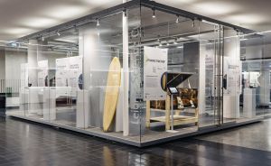Glaswürfel-Ausstellungsraum mit Surfbrett, goldenem Klavier und Informationstafeln in einem modernen Museumsgebäude.