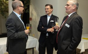 Drei Männer in dunklen Anzügen stehen zusammen und halten Sektgläser bei einer Veranstaltung.