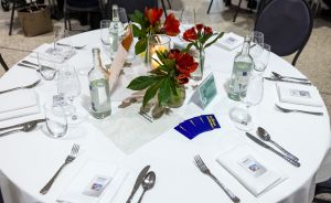 Runder Tisch mit weißer Tischdecke, Besteck, Gläsern, Wasserflaschen, roten Blumen und blauen Karten.