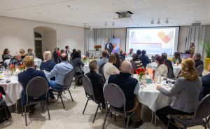 Konferenzraum mit Rundtischen und Publikum, das einem Redner vor einer Präsentationsleinwand zuhört.
