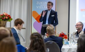 Mann im Anzug spricht vor Publikum bei Veranstaltung, im Hintergrund Banner mit Aufschrift „Zukunft braucht Vielfalt".