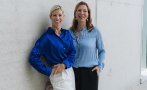Zwei lächelnde Frauen lehnen an einer Betonwand. Eine trägt blaues Seidenhemd, die andere hellblaue Bluse.