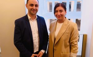 Zwei Personen stehen nebeneinander in einem hellen Gebäude mit Säulen. Mann im Blazer, Frau im Beige-Jacket.