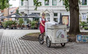 Person mit roter Schürze schiebt Lastenrad mit Aufschrift „Birne & Beere" auf einem Kopfsteinpflasterplatz.