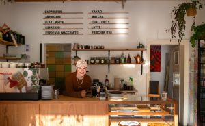 Café-Tresen mit Menütafel, Espressomaschine und Glasvitrine. Eine Person mit Kappe steht lächelnd dahinter.