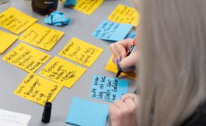 Person schreibt mit Marker auf blaue Haftnotiz; viele beschriftete gelbe und blaue Zettel liegen auf grauem Tisch.