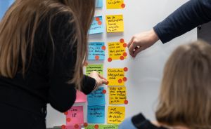 Drei Personen ordnen beschriftete bunte Haftnotizen und rote Klebepunkte an einer weißen Wand an.