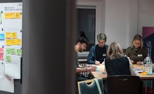 Vier Personen sitzen an einem Tisch und arbeiten mit bunten Haftnotizen. Im Hintergrund hängt ein Whiteboard mit Zetteln.