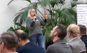 Frau mit Mikrofon spricht vor Publikum, zeigt auf Flipchart mit deutschen Notizen, Palmen im Hintergrund.
