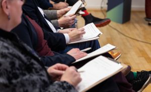 Mehrere Personen sitzen nebeneinander und halten Klemmbrett mit Papieren auf dem Schoß, einige halten Stifte.