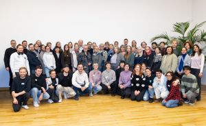Große Gruppe von etwa 50 Personen posiert für ein Gruppenfoto in einem hellen Raum mit Holzboden.