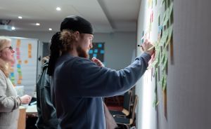 Mann mit Bart und Mütze schreibt mit Marker auf Post-its an einer Wand. Zwei Personen stehen daneben.