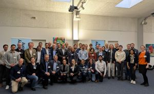 Große Gruppe von etwa 40 Personen posiert für ein Gruppenfoto in einem Konferenzraum mit blauen Aufstellern.
