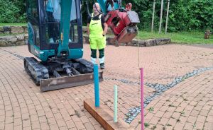 Minibagger auf Pflasterplatz, Arbeiter in Warnkleidung daneben, im Vordergrund ein Holzbalken mit farbigen Stäben.