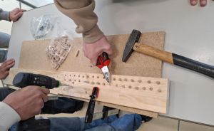 Zwei Personen arbeiten an einem Holzbrett mit Bohrmaschine, Hammer und Zange; Holzdübel und Schrauben liegen daneben.