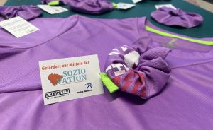 Lila T-Shirts auf grüner Schneidematte mit Haargummis und Karte „Gefördert aus Mitteln des SOZIO NATION, KreHtiv, Region Hannover".