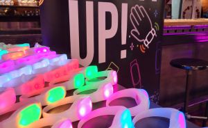 Leuchtende LED-Armbänder in Pink, Grün und Blau vor einem "Light Up!"-Banner in einem Veranstaltungsraum.