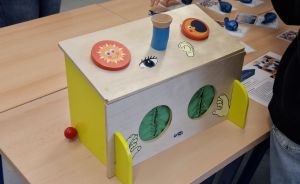 Holzbox mit aufgemalten Sinnesorganen (Auge, Ohr, Hände) und rundem Sonnen-Kreis auf einem Tisch.
