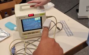 Person mit Pulsoximeter am Finger vor einem medizinischen Monitor mit EKG-Kurve; zweite Hand berührt das Gerät.