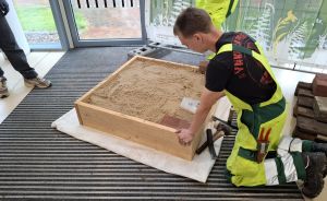 Mann in Warnschutzkleidung kniet vor einer mit Sand gefüllten Holzkiste auf dem Boden eines Gebäudes.