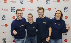 Vier Personen in dunkelblauen Sweatshirts posieren lächelnd vor einem Sparkasse-Hannover-Banner.