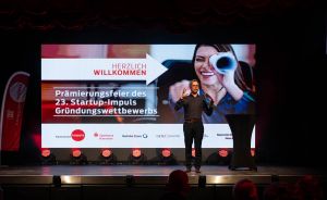 Mann steht auf Bühne vor Leinwand mit Aufschrift „Prämierungsfeier des 23. Startup-Impuls Gründungswettbewerbs".