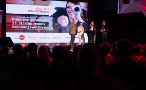 Bühnenszene der Prämierungsfeier des 23. Startup-Impuls Gründungswettbewerbs mit vier Personen vor einer Leinwand.