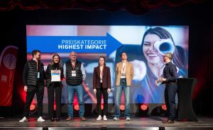 Sechs Personen auf einer Bühne vor einer Leinwand mit der Aufschrift „Preiskategorie Highest Impact".
