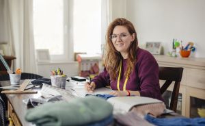 Frau mit roten Haaren, Brille und gelbem Maßband sitzt an einem Schreibtisch mit Stoffen und Stiften.