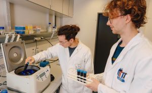 Zwei Personen in weißen Laborkitteln arbeiten mit einem Zentrifugiergerät und halten Reagenzgläser in einem Labor.