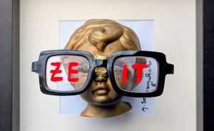 Goldene Kinderkopf-Büste mit schwarzer Brille, auf deren Gläsern „ZEIT" in roten Buchstaben steht, gerahmt.