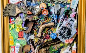 Goldgerahmte Assemblage-Kunst: bunte Cereal-Ringe, Besteck, Pinsel, Flaschen, Perlen, Federn und Alltagsobjekte dicht arrangiert.