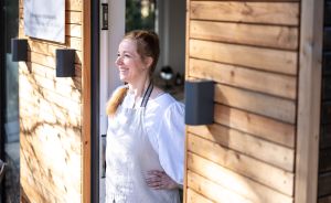 Frau mit rotem Haar und weißer Schürze steht lächelnd in einer Holzverkleideten Eingangstür im Sonnenlicht.