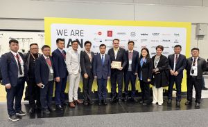 Gruppe von ca. 13 Personen in Businesskleidung posiert vor gelbem Banner mit Aufschrift „WE ARE" bei einer Messe.