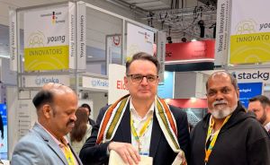 Drei Männer stehen an einem Stehtisch auf einer Messe. Der mittlere trägt einen Schal und hält ein Buch.