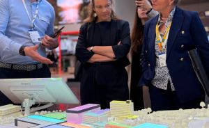 Drei Personen betrachten ein beleuchtetes Stadtmodell auf einer Messe. Ein Mann mit Badge erklärt, zwei Frauen hören zu.