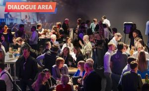 Viele Menschen stehen und sitzen in einem Veranstaltungsraum mit lila Beleuchtung unter einem Schild "Dialogue Area".