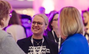 Frau mit Brille und schwarzem T-Shirt „Machen Einfach Machen" lächelt bei Veranstaltung, trägt Konferenz-Badge.