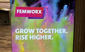 Leuchtende Werbetafel von „FEMWORX" mit dem Slogan „Grow together. Rise higher." und buntem Farbpulver-Hintergrund.