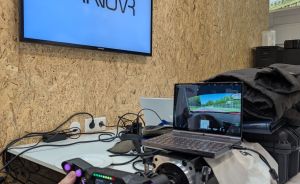 Person hält Simracing-Lenkrad mit lila Knöpfen. Im Hintergrund Laptop mit Fahrsimulation und TV mit "HANOVR"-Logo.