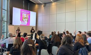 Publikum mit erhobenen Händen in einem modernen Konferenzraum; zwei Personen stehen vor einer Präsentationsleinwand.