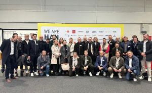 Gruppe von ca. 30 Personen posiert vor gelbem Banner mit Aufschrift „We Are HAN" in einer Messehalle.