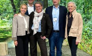 Fünf Personen stehen lächelnd vor einer Fototapete mit Waldmotiv auf einem gepflasterten Boden.