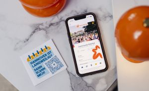 Smartphone zeigt App mit "Community Picknick", daneben Flyer mit QR-Code und Text "Hannover ist langweilig? Dann scann mich!"