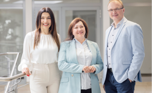 Drei Personen stehen lächelnd nebeneinander in einem hellen Gebäude, zwei Frauen und ein Mann in Business-Kleidung.