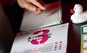 Hand mit Stift über Notizbuch, davor Flyer mit Logo „Fireabend x Hannovr", daneben weiße Gummiente auf dunklem Tisch.
