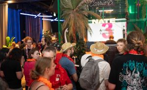 Menschengruppe auf einer Party mit Strohhüten und orangefarbenen Blumenketten, tropischer Dekoration und Neonschild „NATE".