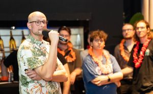Ein kahlköpfiger Mann mit Brille spricht in ein Mikrofon. Im Hintergrund stehen mehrere Personen mit orangefarbenen Blumenketten.